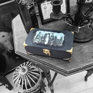 May include: A rectangular black wooden box featuring gold-toned metal corner protectors and a central gold latch. The lid displays a monochromatic graphic print of several horned figures. The box has a dark finish with visible texture.