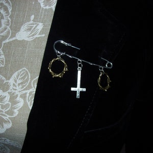 Puede incluir: Un broche de seguridad plateado con tres dijes: una cruz invertida dorada y dos anillos dorados con diseño de espinas.