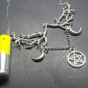 May include: Silver-toned necklace featuring a yellow and silver battery pendant, a branch-like design, crescent moons, and a pentagram charm. The necklace has a chain and is set against a dark background.