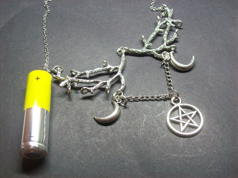 May include: Silver-toned necklace featuring a yellow and silver battery pendant, a branch-like design, crescent moons, and a pentagram charm. The necklace has a chain and is set against a dark background.