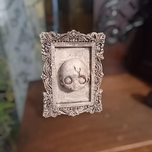 Könnte beinhalten: Eine kleine, weiße, gerahmte Skulptur eines Babykopfes mit großen, dunklen Augen. Der Rahmen ist verziert und hat eine abgenutzte Oberfläche.