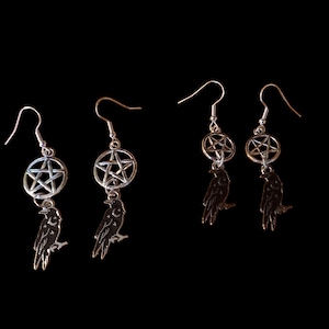 Puede incluir: Pendientes plateados con un pentagrama y un colgante de cuervo. Los pendientes tienen cierre de gancho y se muestran sobre un fondo negro. El cuervo está posado en una rama.