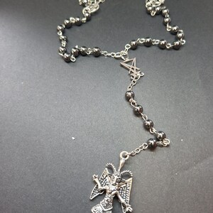 K&ouml;nnte beinhalten: Eine Halskette mit dunklen, metallischen, kugelf&ouml;rmigen Perlen an einer silberfarbenen Kette. Das Schmuckst&uuml;ck enth&auml;lt einen silberfarbenen Anh&auml;nger in Form einer gefl&uuml;gelten Figur mit H&ouml;rnern und ein zentrales silberfarbenes Symbol.