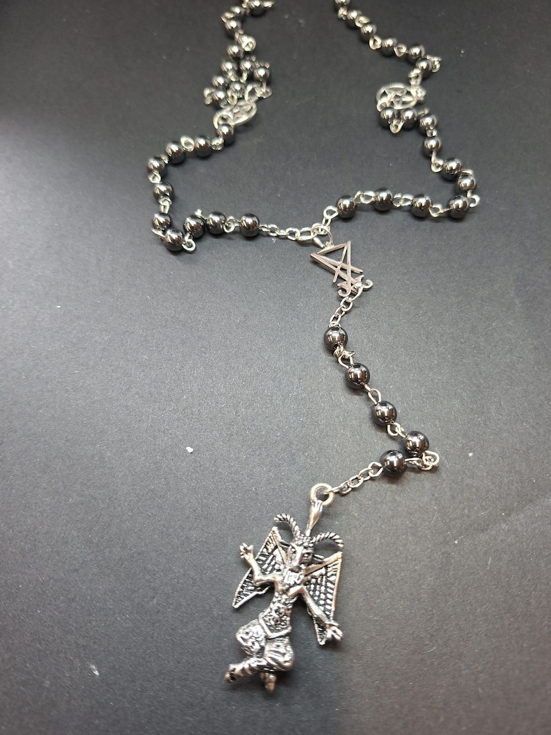 K&ouml;nnte beinhalten: Eine Halskette mit dunklen, metallischen, kugelf&ouml;rmigen Perlen an einer silberfarbenen Kette. Das Schmuckst&uuml;ck enth&auml;lt einen silberfarbenen Anh&auml;nger in Form einer gefl&uuml;gelten Figur mit H&ouml;rnern und ein zentrales silberfarbenes Symbol.