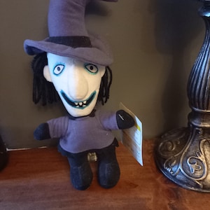 May include: A plush toy of the character Oogie Boogie from the movie The Nightmare Before Christmas. The toy is wearing a purple hat and a purple dress. The toy has a white face with a large nose and a wide smile.