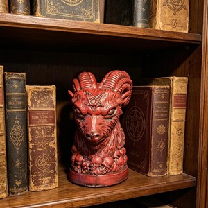 May include: A red sculpture of a ram head with a pentagram symbol on its forehead, positioned on a base decorated with small skulls. The ram features textured, curved horns and detailed facial features, standing between several antique books.