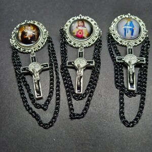 Op de afbeelding: Drie religieuze broches met gedetailleerde zilverkleurige frames en zwarte kettingen. Elke broche heeft een andere afbeelding in een cirkelvormig frame, met een kruisvormige hanger eronder. De kruisen hebben een zilverkleurig kruisbeeld en een zwarte achtergrond.