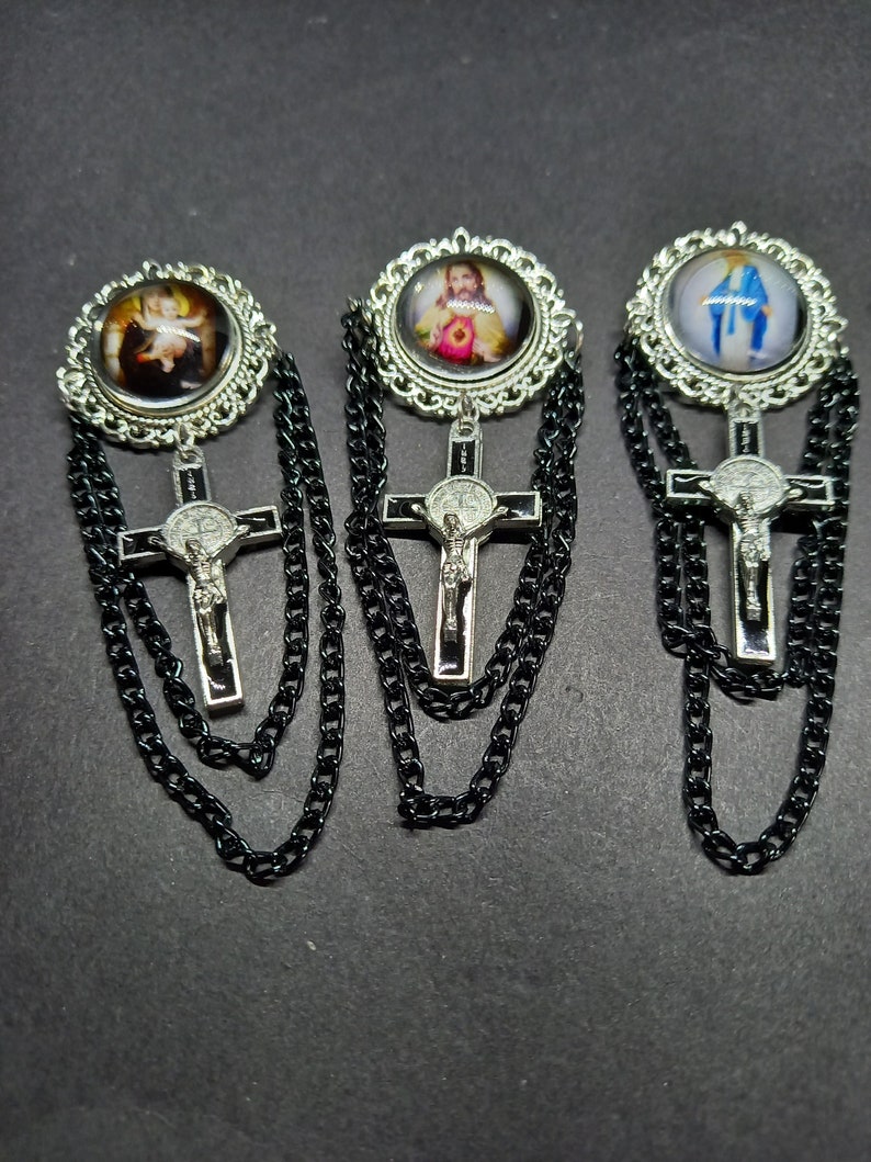 Op de afbeelding: Drie religieuze broches met gedetailleerde zilverkleurige frames en zwarte kettingen. Elke broche heeft een andere afbeelding in een cirkelvormig frame, met een kruisvormige hanger eronder. De kruisen hebben een zilverkleurig kruisbeeld en een zwarte achtergrond.