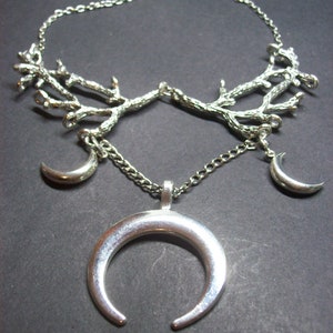 May include: Silver-toned necklace featuring a branch-like design at the top, with three crescent moon pendants. The central pendant is a large, open crescent moon. The necklace is on a dark background.
