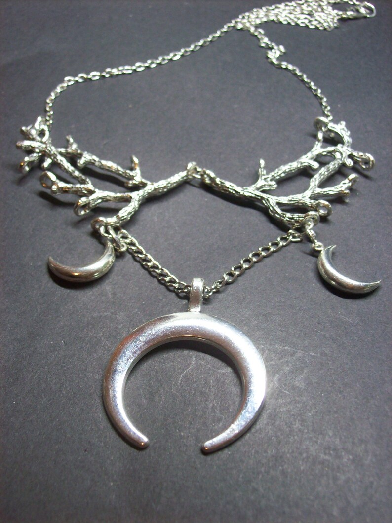 May include: Silver-toned necklace featuring a branch-like design at the top, with three crescent moon pendants. The central pendant is a large, open crescent moon. The necklace is on a dark background.