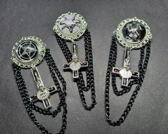 Op de afbeelding: Drie zilverkleurige broches met ronde frames en zwart-witte grafische inzetstukken. Elke broche bevat een hangende omgekeerde crucifix-hanger en twee zwarte metalen kettingen die het frame met het kruis verbinden, wat zorgt voor een gelaagd ontwerp.