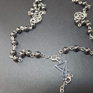 K&ouml;nnte beinhalten: Eine Halskette mit dunkelmetallischen, kugelf&ouml;rmigen Perlen, die durch silberfarbene Metallketten verbunden sind. Das Design umfasst zwei kreisf&ouml;rmige Pentagramm-Anh&auml;nger und ein zentrales, silberfarbenes Sigillen-Symbol.