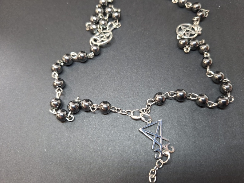 K&ouml;nnte beinhalten: Eine Halskette mit dunkelmetallischen, kugelf&ouml;rmigen Perlen, die durch silberfarbene Metallketten verbunden sind. Das Design umfasst zwei kreisf&ouml;rmige Pentagramm-Anh&auml;nger und ein zentrales, silberfarbenes Sigillen-Symbol.