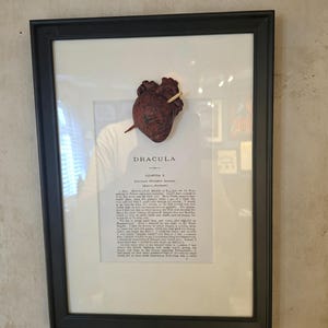 Könnte beinhalten: Ein gerahmtes Wandkunstwerk mit einer Seite aus dem Buch Dracula, Kapitel 1, mit einer dreidimensionalen, dunkelroten anatomischen Herzskulptur, die in der Mitte des Papiers montiert und von einem kleinen hellen Holzpflock durchbohrt ist.