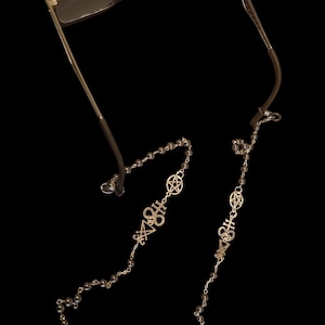 May include: Dark-framed eyeglasses with a silver chain featuring occult symbols and small beads. The chain is designed to hold the glasses and has a decorative, gothic aesthetic. The background is black.