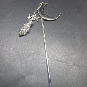 Może przedstawiać: Srebrzysta metalowa spinka do włosów z dużym półksiężycem u góry. Zawieszka przymocowana łańcuszkiem zawiera mały pentagram otoczony dwoma maleńkimi półksiężycami oraz przezroczysty kryształ owinięty drutem, który zwisa poniżej.