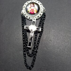 Op de afbeelding: Zilverkleurige broche met een religieuze afbeelding in een decoratief frame. Een zilveren kruis met een zwart midden hangt eronder, met zwarte kettingaccenten. De broche heeft een vintage of gotische uitstraling.