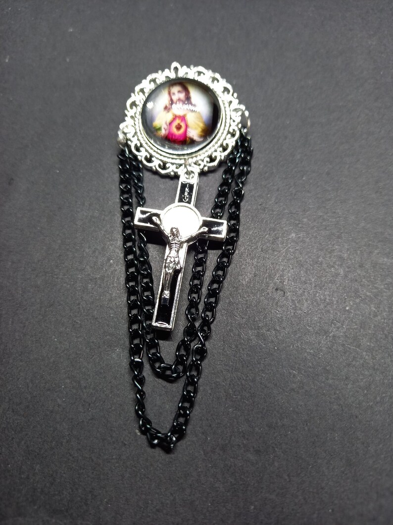 Op de afbeelding: Zilverkleurige broche met een religieuze afbeelding in een decoratief frame. Een zilveren kruis met een zwart midden hangt eronder, met zwarte kettingaccenten. De broche heeft een vintage of gotische uitstraling.