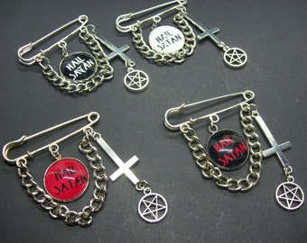 Op de afbeelding: Vier zilveren veiligheidsspeld-broches met een ketting, een kruis en een pentagram-bedel. Elke broche heeft een ronde knoop in zwart-wit of rood-zwart met de tekst "Hail Satan".