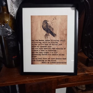 May include: Framed artwork featuring a detailed illustration of a raven perched on a branch, with text from a poem below. The artwork is in a black frame and has a vintage, aged paper appearance.