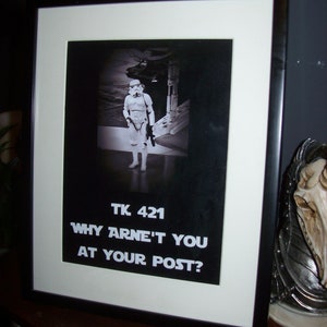 May include: A black and white framed photograph of a Star Wars Stormtrooper standing in a dark corridor. The photo has the text "TK 421 Why aren't you at your post?" printed below the image.