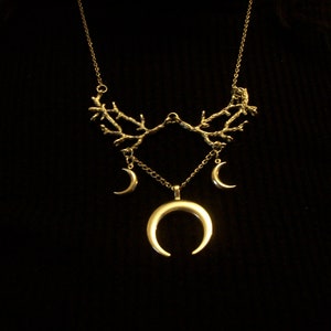 May include: A gold-colored necklace with a celestial theme. The necklace features a branch-like design at the top, with three crescent moon charms hanging below. The largest crescent moon is at the center.