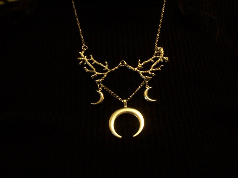 May include: A gold-colored necklace with a celestial theme. The necklace features a branch-like design at the top, with three crescent moon charms hanging below. The largest crescent moon is at the center.