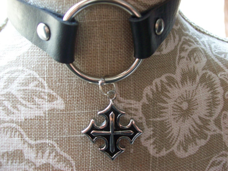 May include: A black leather choker with a silver ring and a silver cross pendant. The cross has a black center.