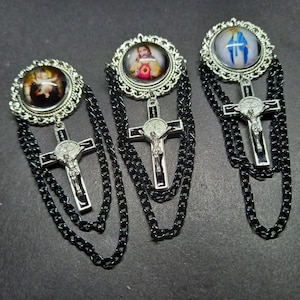 Op de afbeelding: Drie religieuze broches met zilveren kruisen en zwarte kettingaccenten. Elke broche heeft een andere afbeelding in een decoratief zilveren frame. De afbeeldingen omvatten een baby-engel, Jezus en de Maagd Maria.