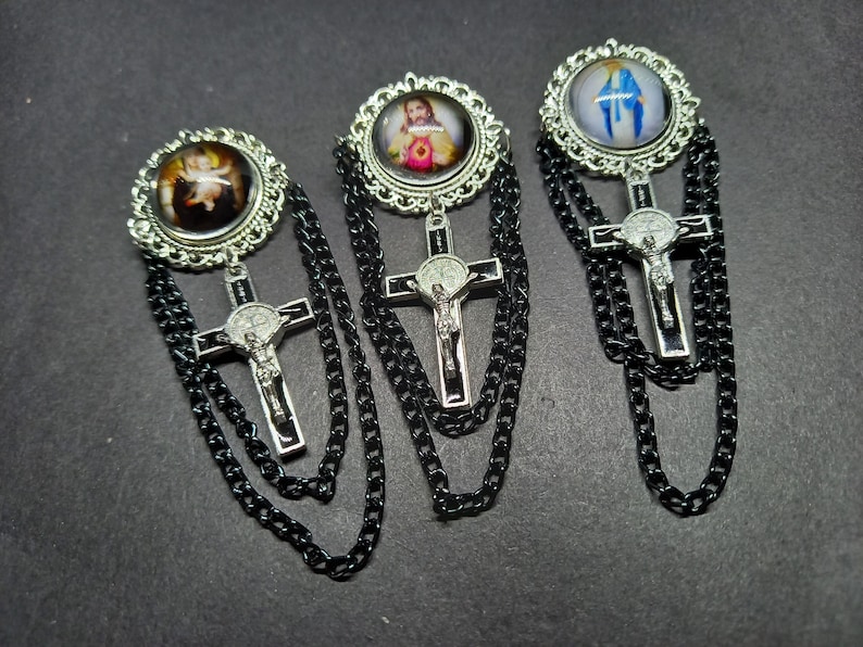 Op de afbeelding: Drie religieuze broches met zilveren kruisen en zwarte kettingaccenten. Elke broche heeft een andere afbeelding in een decoratief zilveren frame. De afbeeldingen omvatten een baby-engel, Jezus en de Maagd Maria.