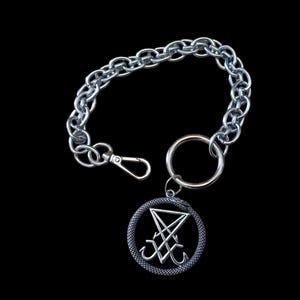 Può includere: Catena color argento con moschettone e un anello grande. Un ciondolo circolare con un simbolo stilizzato pende dall'anello. Il ciondolo ha un bordo a forma di serpente e un design centrale.