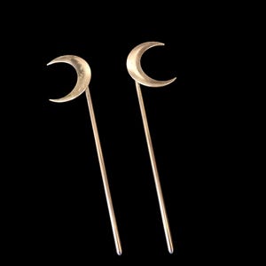 May include: Two matching hair sticks crafted from a metallic material, each featuring a smooth, polished crescent moon topper attached to a long, slender, straight rod. The design is uniform, with both pieces displaying a reflective, light gold finish.