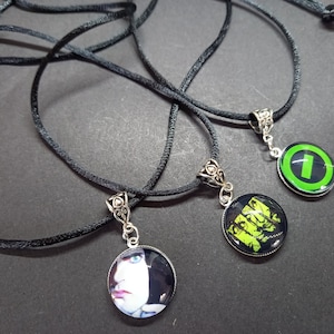 May include: Three black cord necklaces with circular pendants. One pendant displays a face, another a green and yellow design, and the third a green number one on a black backdrop. Each pendant has a silver-coloured decorative bail.