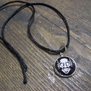 May include: A black cord necklace with a round silver pendant featuring a white skull with a toothy grin and large eyes.