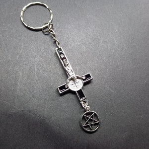 Może przedstawiać: Srebrny brelok do kluczy z czarnym odwróconym krzyżem i srebrnym pentagramem. Krzyż przedstawia srebrną postać z aureolą.
