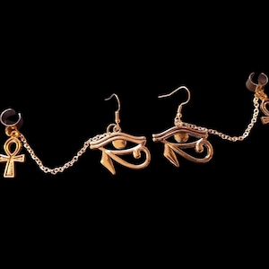 以下が含まれることがあります： 金色のエジプトをテーマにしたイヤリング。片方のイヤリングはフックとホルスの目のデザインで、もう片方はアンクのシンボルと黒いイヤーカフに繋がれたチェーンが付いています。イヤリングは黒い背景にあります。