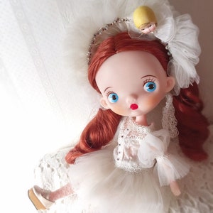 May include: A red-haired doll wearing a white tulle dress with lace detailing. The doll has blue eyes and red lips. She is wearing a white headband with a yellow flower on top. The doll is sitting on a white lace surface.