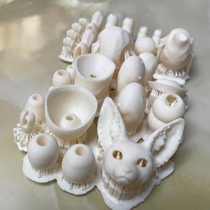 May include: A white 3D printed sculpture of a cat's head with large ears and a detailed face. The sculpture is surrounded by other smaller 3D printed objects, including spheres, cups, and rings.