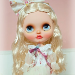 Può includere: Una bambola con lunghi capelli biondi, che indossa un vestito bianco con stampa floreale e un nastro rosa. La bambola ha gli occhi azzurri e le labbra rosa.