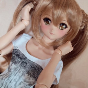 May include: A doll with long, light brown hair styled in pigtails. The doll is wearing a white t-shirt with a black and white graphic print. The doll has blue eyes and rosy cheeks.
