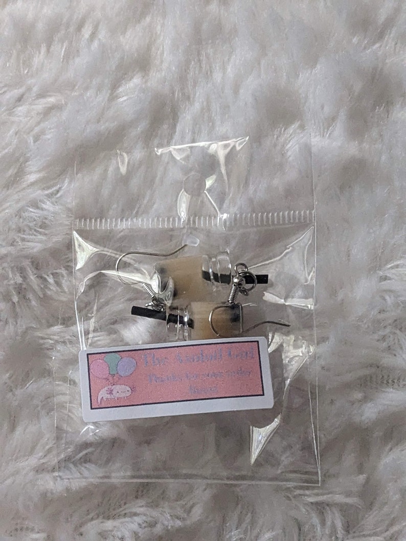Puede incluir: Un par de pendientes de plata con un peque&ntilde;o dije blanco texturizado. Los pendientes est&aacute;n empaquetados en una bolsa de pl&aacute;stico transparente con una peque&ntilde;a etiqueta rosa y blanca que dice "The Axoloti Shop" y "Axoloti, para tu magia interior".