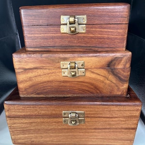 May include: A stack of three rectangular wooden boxes with a natural brown grain finish. Each box features a hinged lid secured by a brass-colored metal latch centered on the front. The boxes are arranged in descending order of size from bottom to top.
