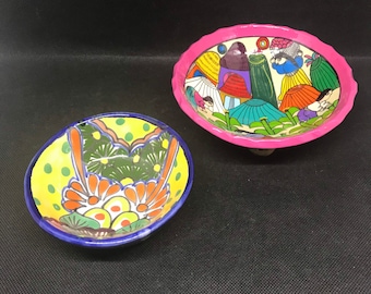 May include: Two small, hand-painted ceramic bowls. One is yellow with green and orange floral designs and a blue rim. The other is oval-shaped with a pink scalloped edge and a painted scene of people with colorful clothing.