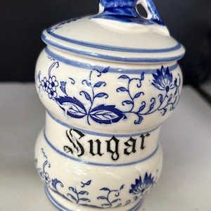 May include: A white ceramic sugar canister featuring a blue floral pattern and the word Sugar printed in black gothic-style lettering. The canister has a rounded body with a matching lid that includes a blue decorative handle on the top.