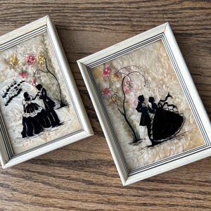 May include: Two framed silhouette art pieces. Each features a black silhouette of a couple dancing under a tree with pink and yellow flowers. The frames are rectangular with a cream-colored border.