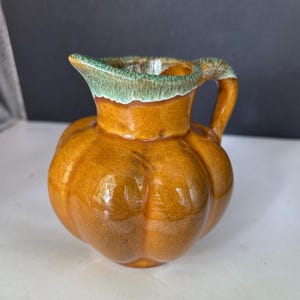 May include: A small ceramic pitcher with a rounded, pumpkin-like shape and a glossy amber-brown glaze. The rim and the top of the handle feature a contrasting drip-style glaze in a muted teal-green color, adding texture to the smooth surface.