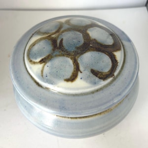 May include: A round, light blue ceramic jar with a lid. The lid features a design of brown, stylized floral shapes on a white background. The jar has a smooth, glossy finish.