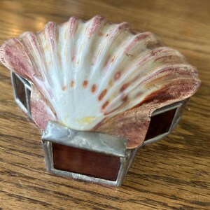 May include: A decorative seashell box with stained glass accents. The shell is a light pink and white color with a brown rim. The stained glass is a dark red color.