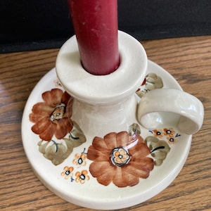May include: A white ceramic candle holder with a brown floral design. The holder has a ring handle and a round base.