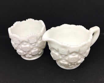 May include: A white milk glass creamer and sugar bowl set with a raised diamond pattern. The creamer has a handle and a pouring spout.